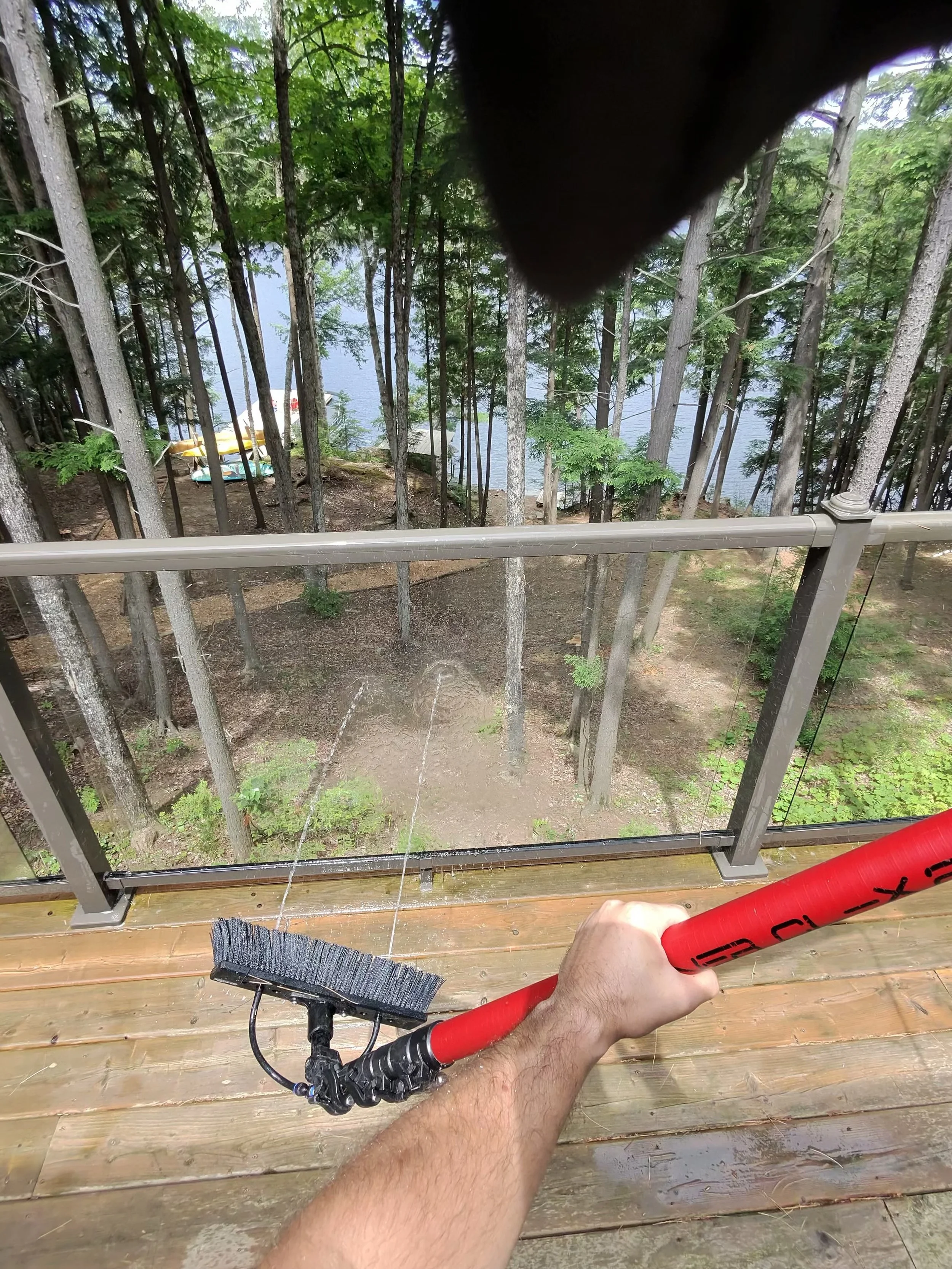 A person using a power washer to clean a wooden deck on a balcony overlooking a wooded area with a lake.