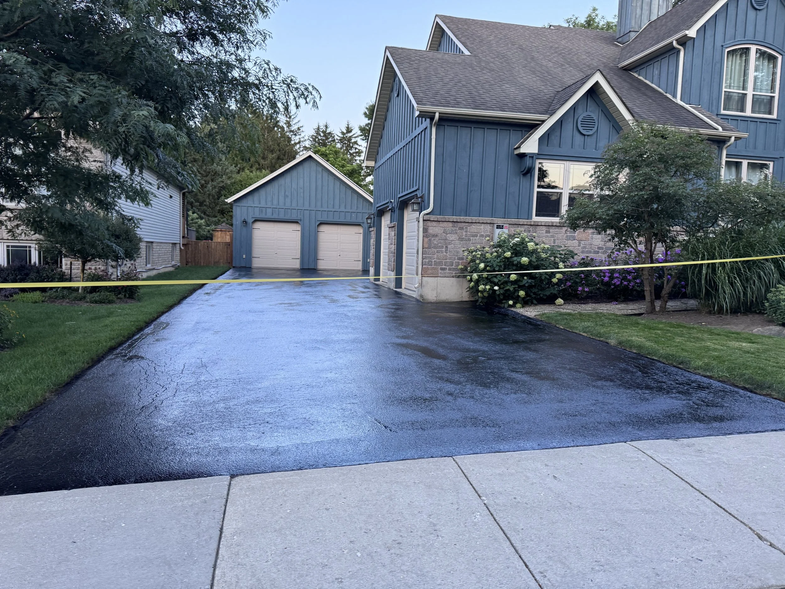 A blue house with a detached garage, wet driveway, and a yellow caution tape across the driveway.