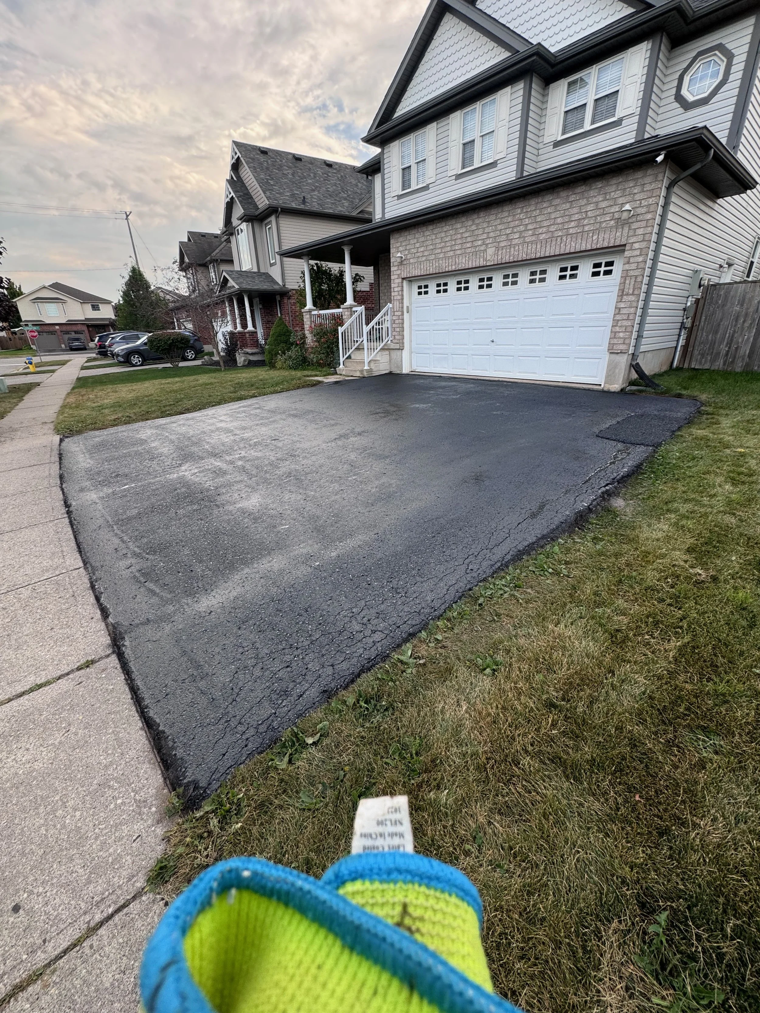 Freshly paved driveway in front of a house, with small grass lawn on item sides, and neighboring houses visible in the background under a cloudy sky.