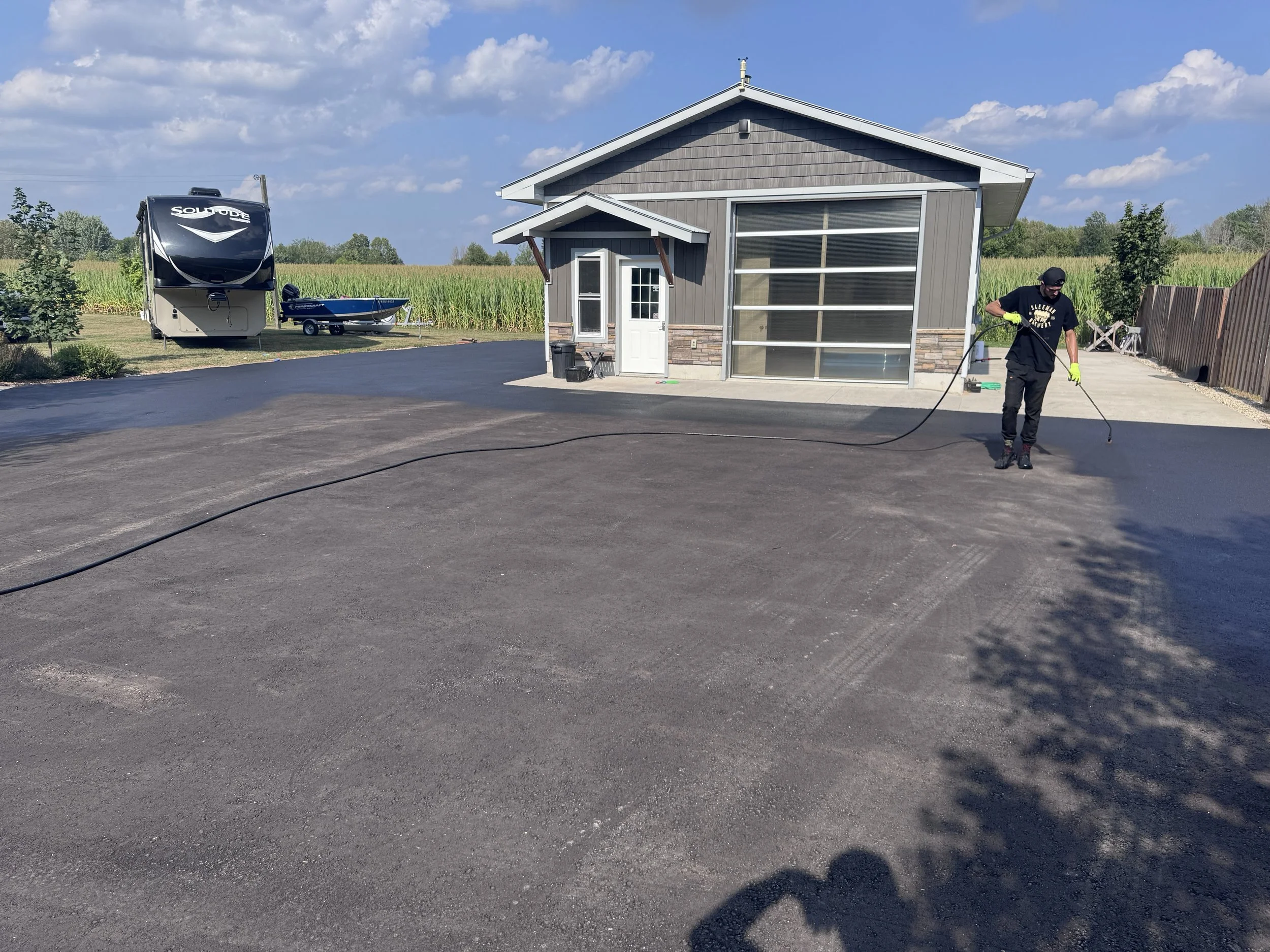 A man wearing gloves is pressure washing a newly asphalt driveway in front of a gray house with a garage. There is a trailer and a boat in the background, along with a field of crops and a partly cloudy sky.