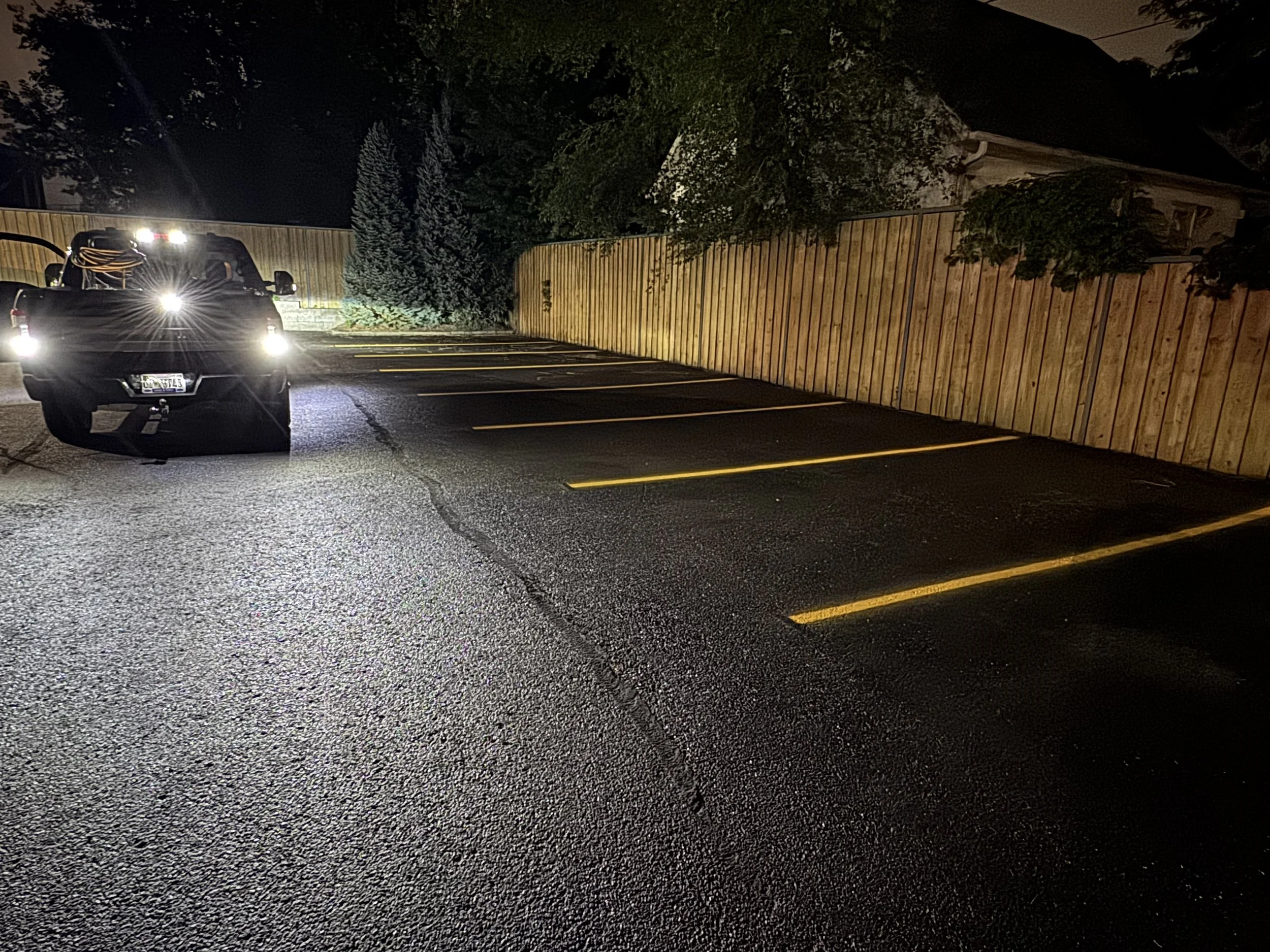 A parking lot at night with an empty row of parking spaces bordered by a wooden fence. A pickup truck with its headlights on is parked at the front, illuminating the area.