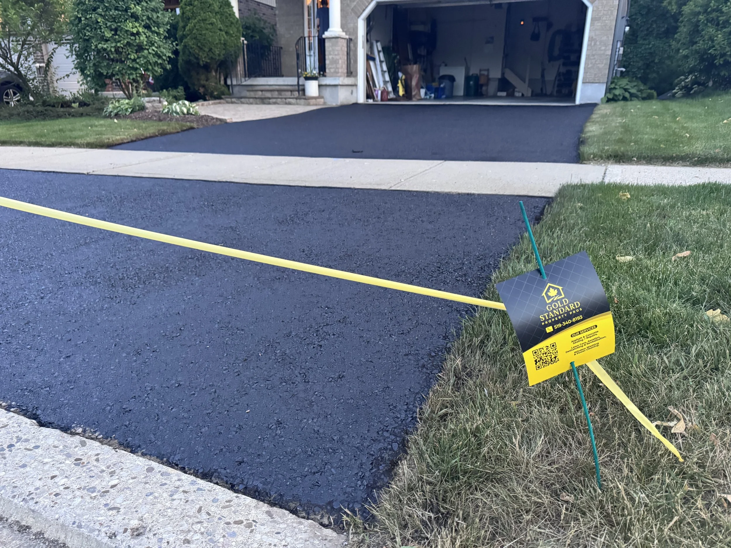 Newly paved black asphalt driveway with a yellow string and a sign indicating it is freshly paved by Gold Standard Paving & Propane.