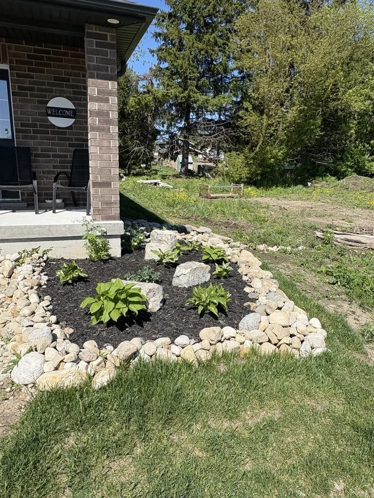 A freshly landscaped flower bed with green plants, surrounded by a border of rocks, located near a house with a brick exterior and a welcome sign on the wall.