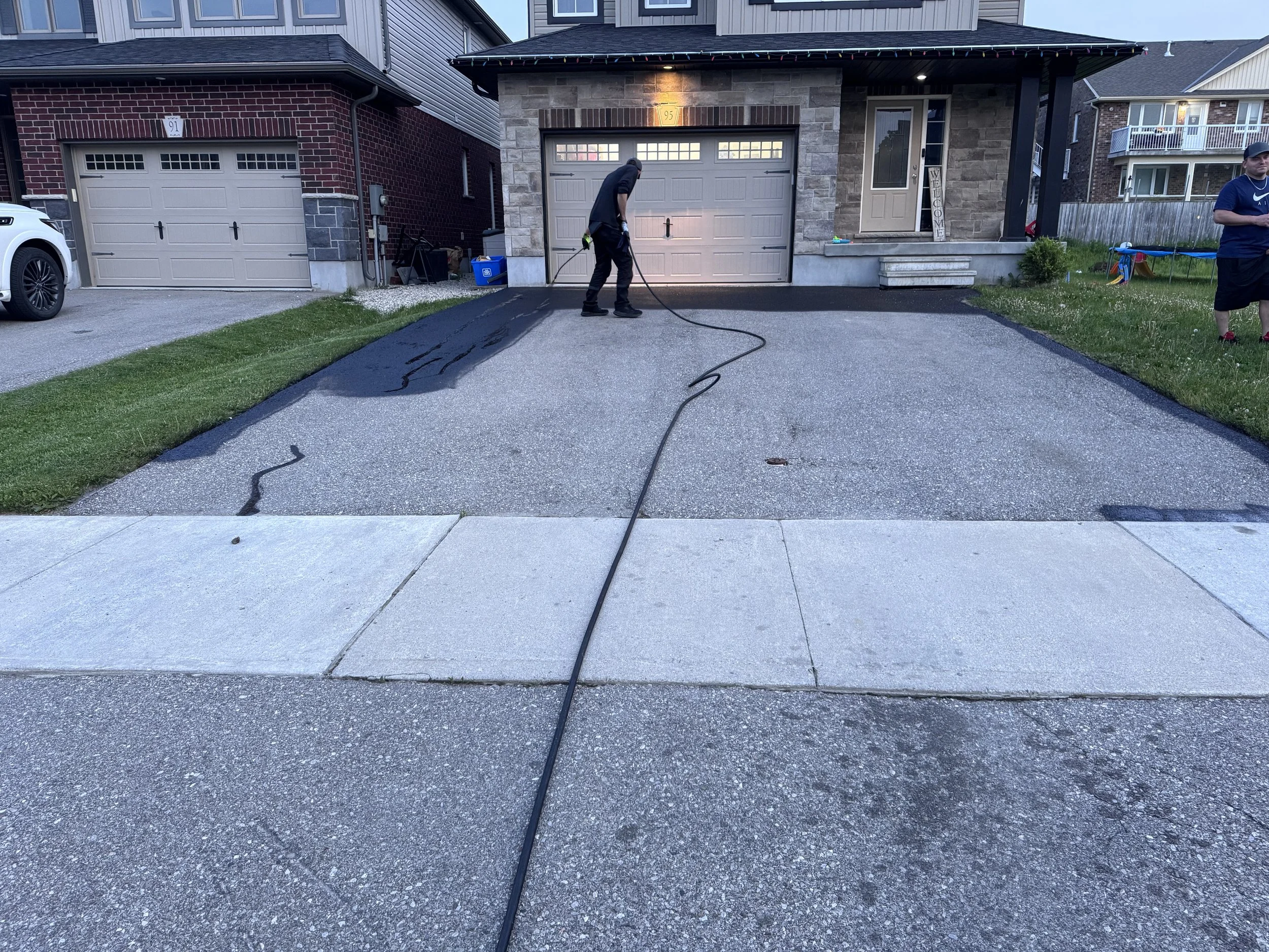 Person pressure washing a driveway in front of a house, with another person standing on the side. The house has a brick and stone exterior, with a garage and a porch.