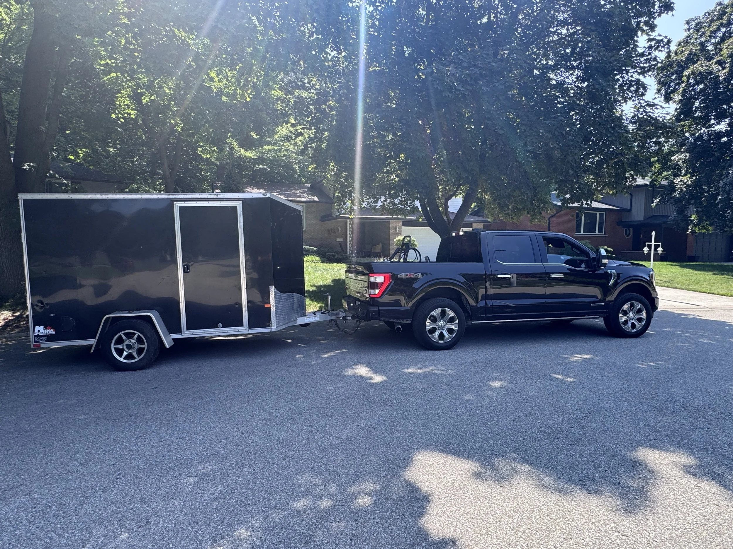 Black pickup truck towing a large black enclosed trailer parked on a suburban street with trees and houses in the background.