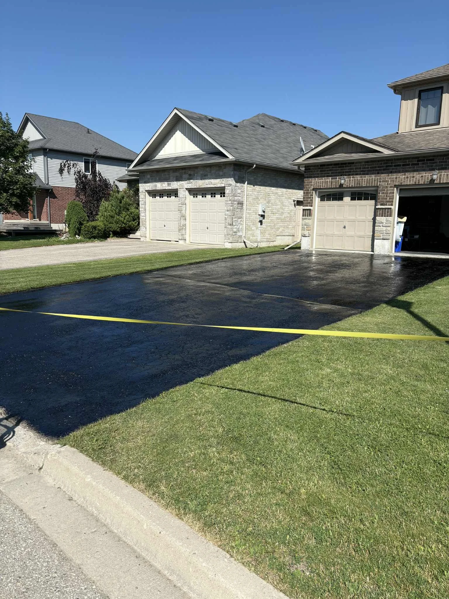 Newly paved black asphalt driveway in front of a residential garage, with green grass on either side, under a clear blue sky.