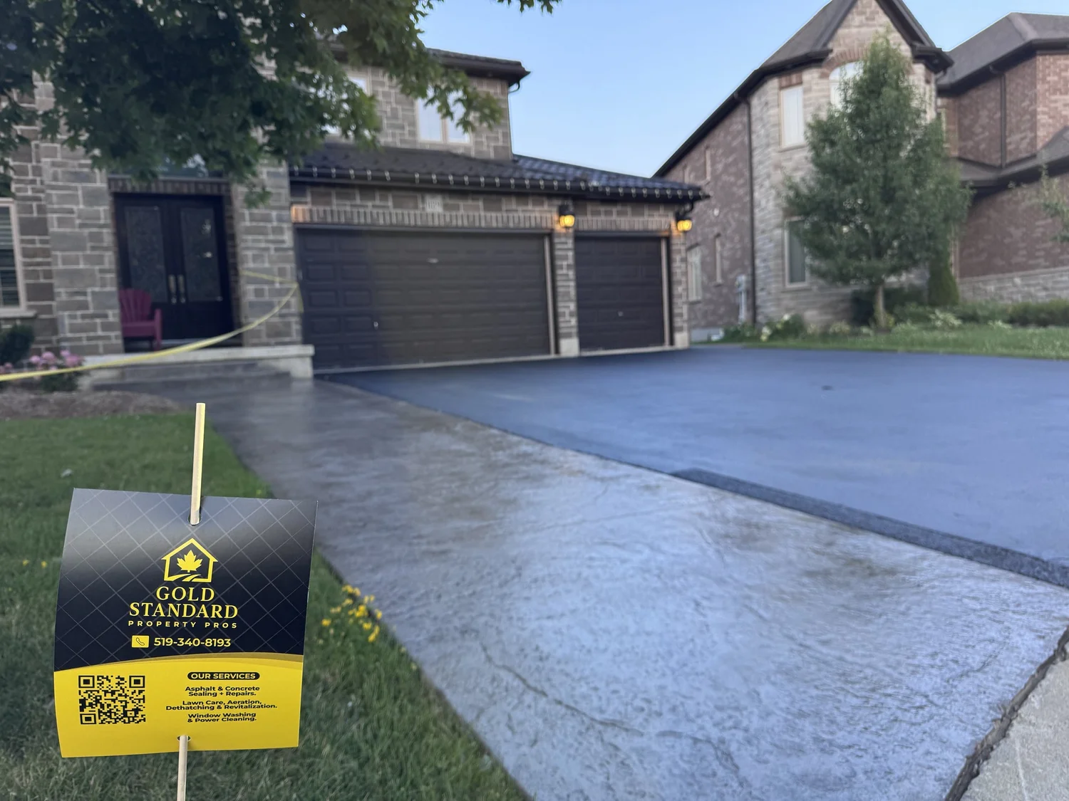 Freshly poured concrete driveway in front of a suburban house with two garages, a tree, and a yellow and black sign for Gold Standard Property Pros in the grass.