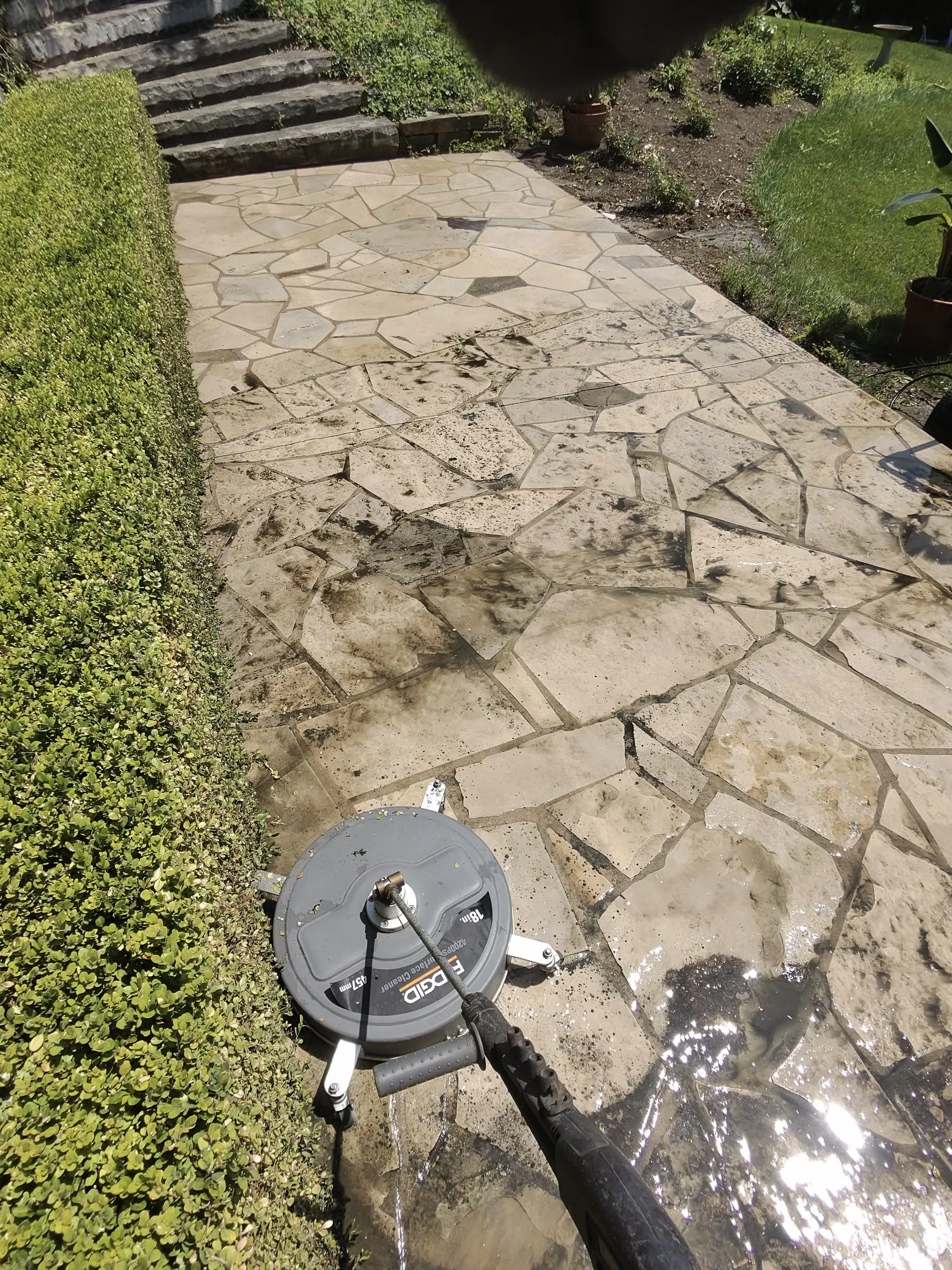 Cleaning a stone patio with a pressure washer near a trimmed hedge and garden steps.