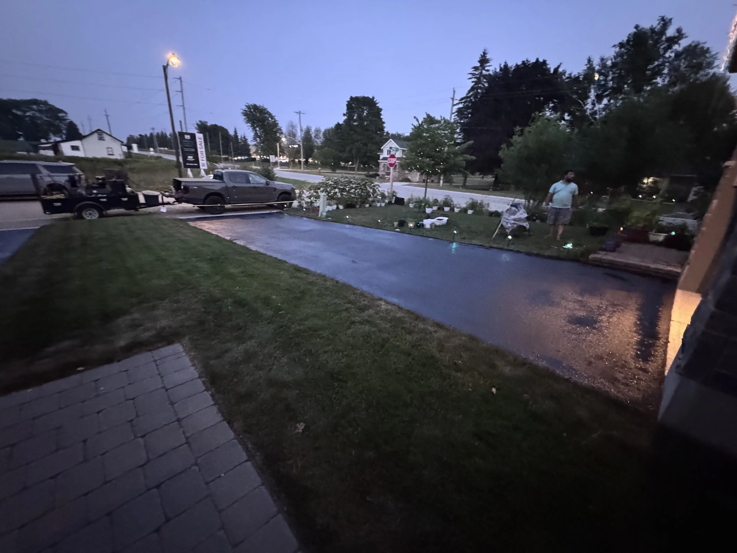 A residential scene at dusk with a wet driveway, a person standing near a flower garden, and vehicles parked along the street.