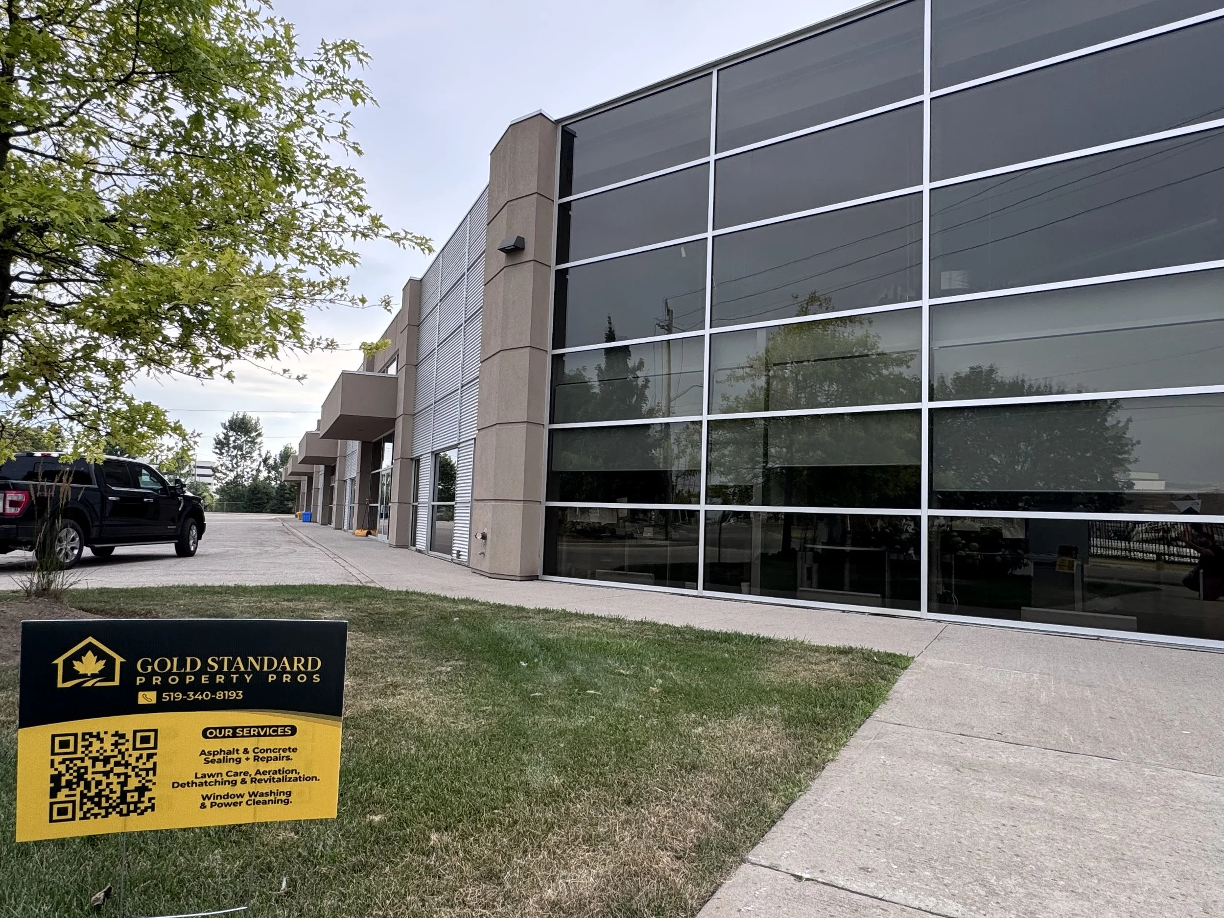 A commercial building with large windows and a sidewalk in front. A black truck is parked on the left side. A sign in the grass advertises Gold Standard Property Pros with services listed, including asphalt, concrete, sealing, repairs, lawn care, and