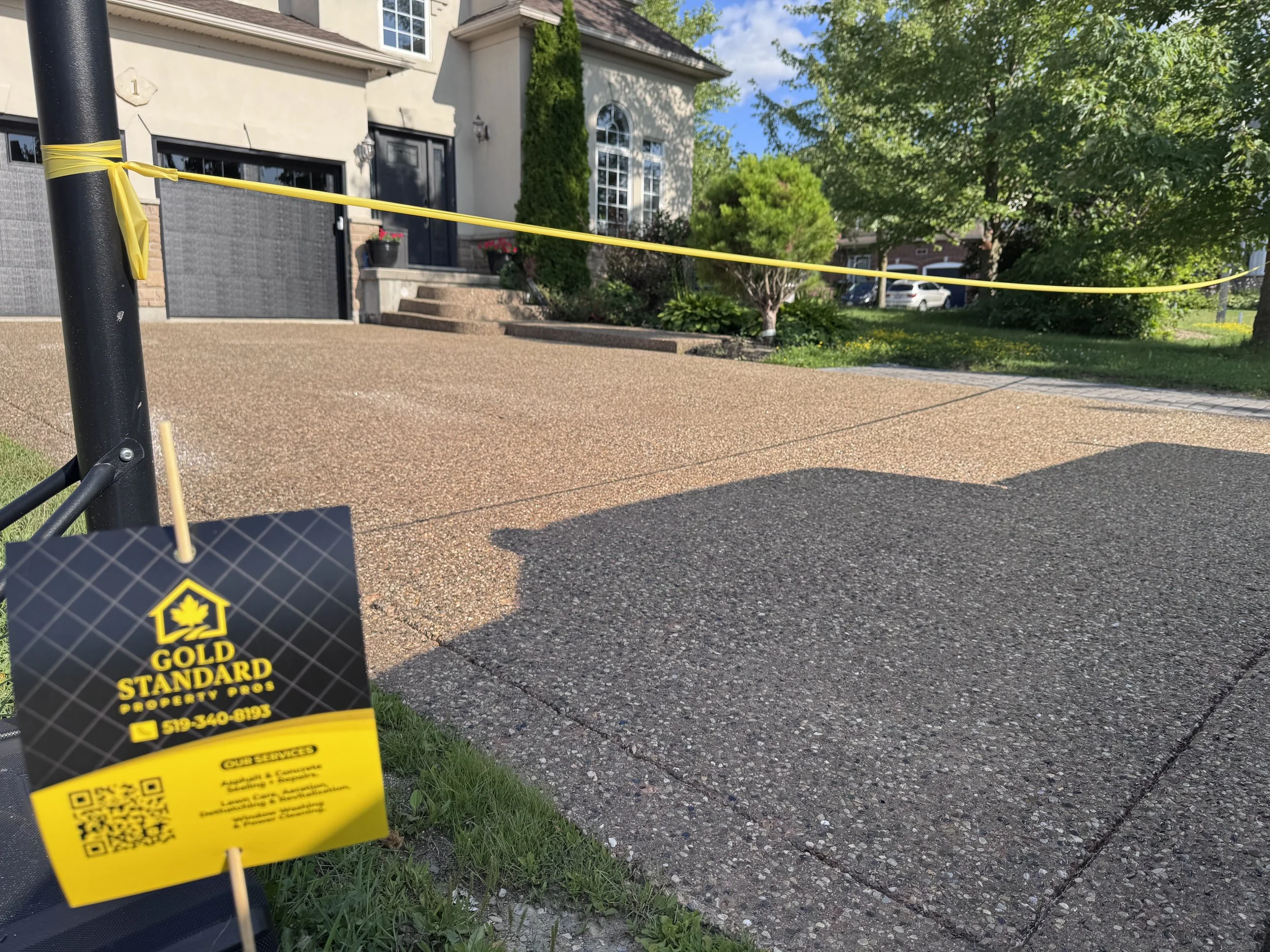A newly poured concrete driveway in front of a house with steps leading to a front door, orange barrier tape, and a black and yellow Gold Standard Property Services sign.