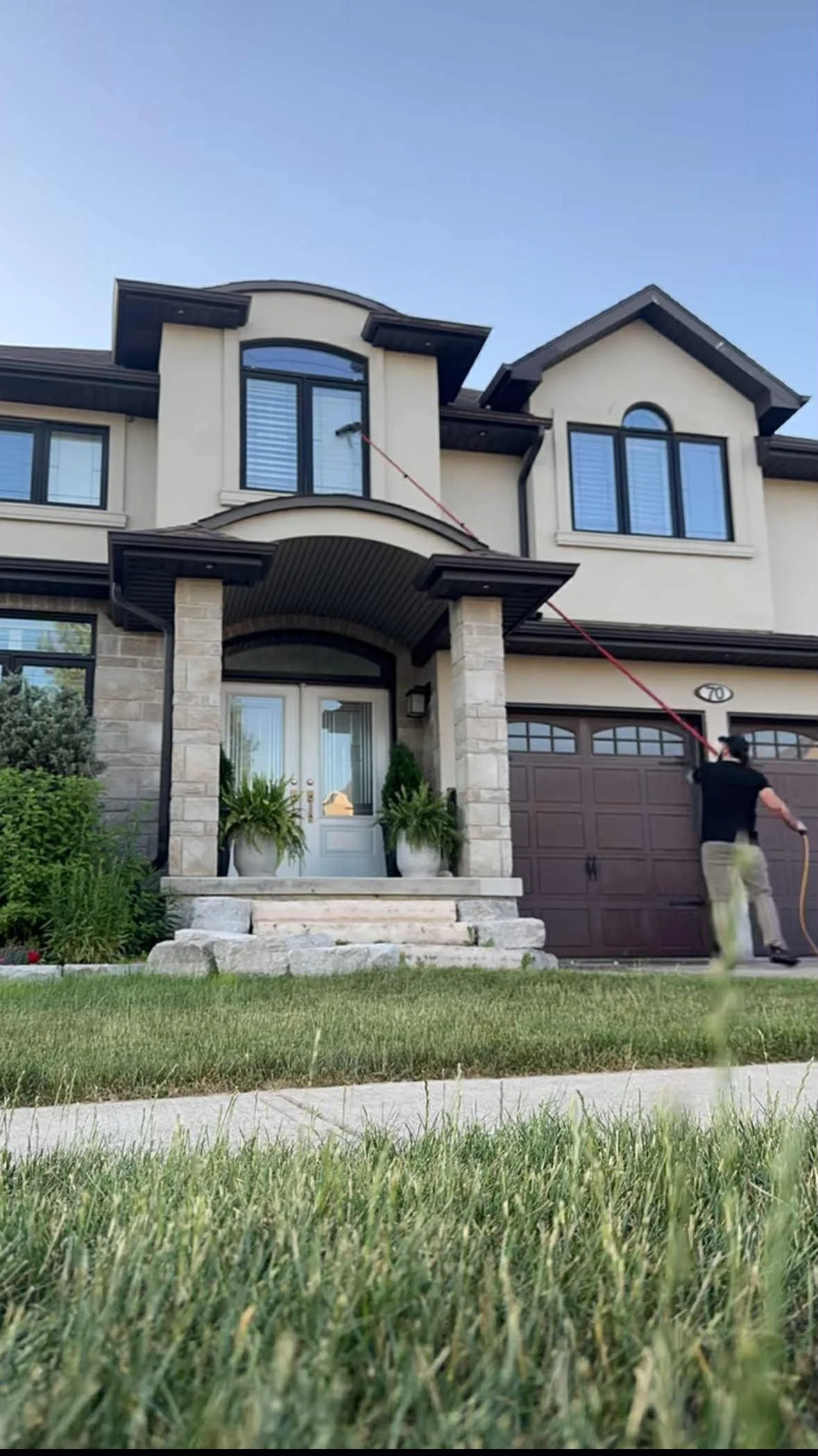 A man in black shirt and khaki pants using a long pole to clean windows of a multi-story beige house with stone accents, located on a green lawn with plants and bushes, under a clear blue sky.