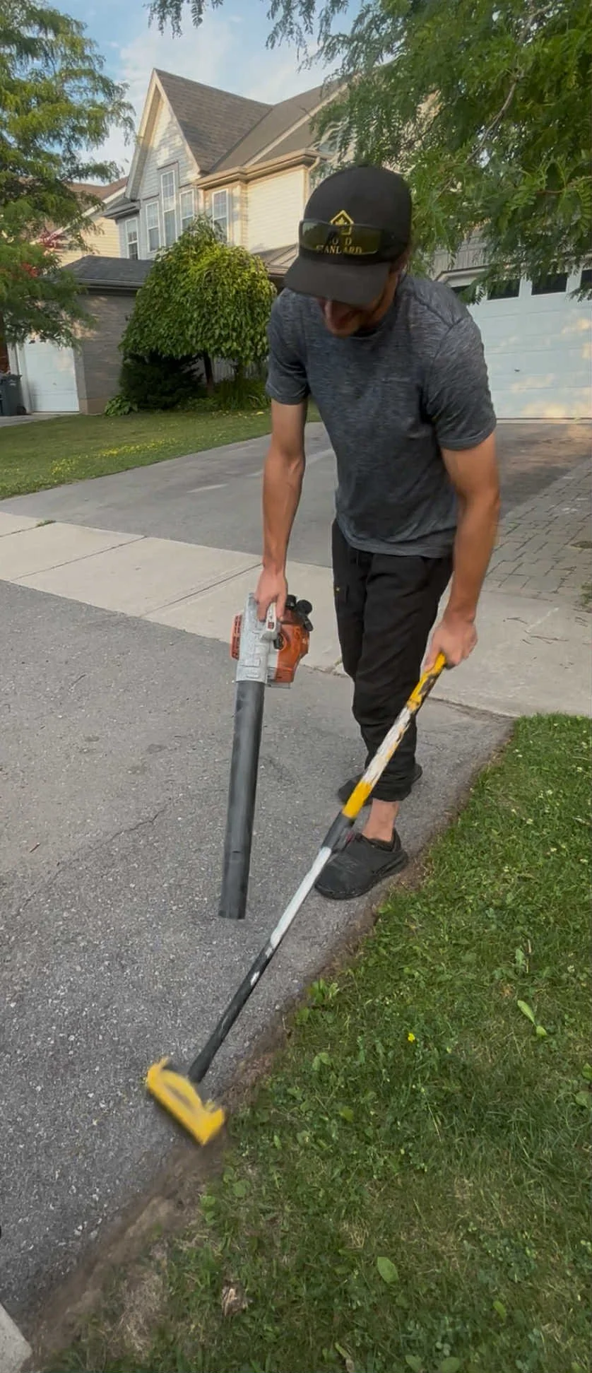 A man using a power edger to trim the edge of a lawn next to a sidewalk in a residential neighborhood.