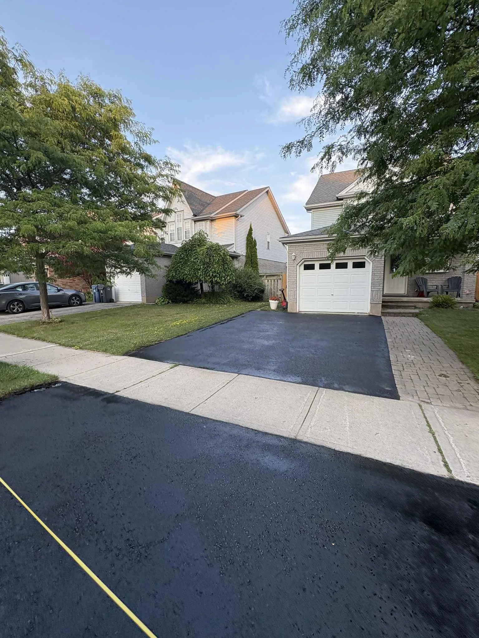 Residential driveway with freshly paved black asphalt, adjacent to the sidewalk, leading to a white garage door of a two-story house. The house has light-colored siding, and the yard has trees, bushes, and a small garden area.