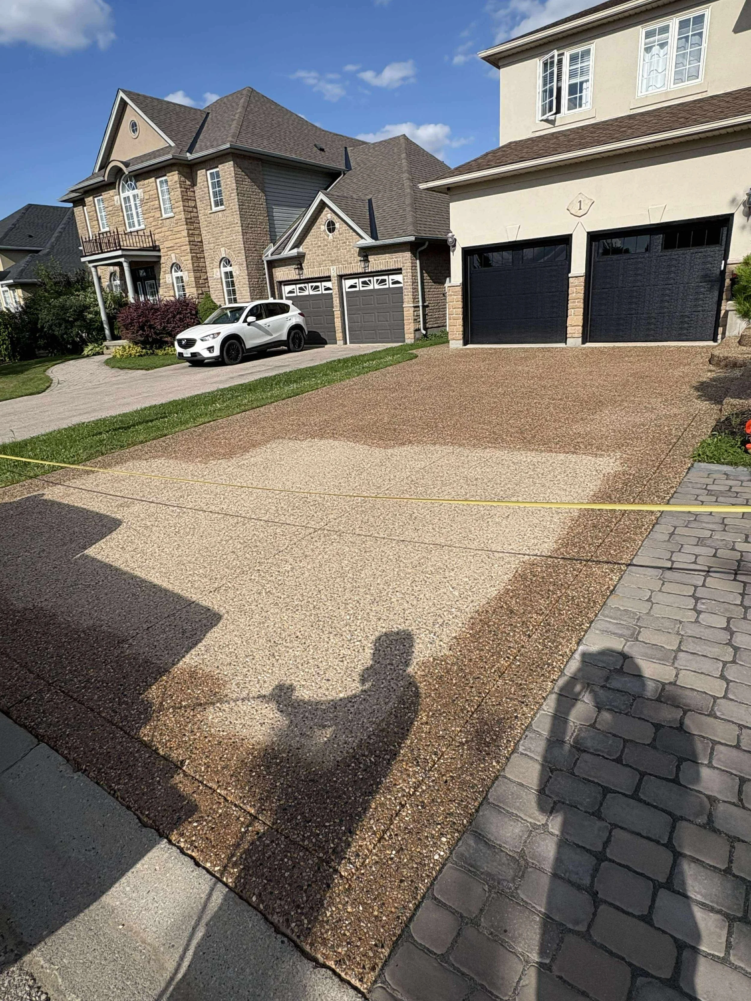 A residential driveway with a textured concrete surface, bordered by a stone walkway and green grass, in front of multi-story houses with brick and siding exteriors, and a white car parked on the driveway. Shadows of a person and a light stand are vi