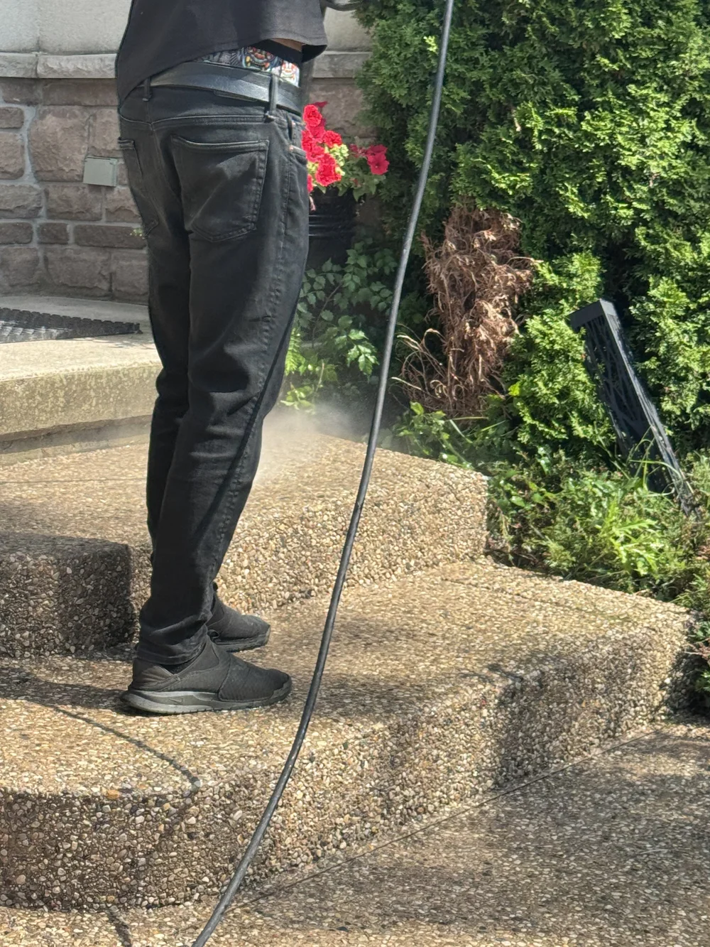 Person pressure washing concrete steps outdoors, with plants and a stone wall in the background.