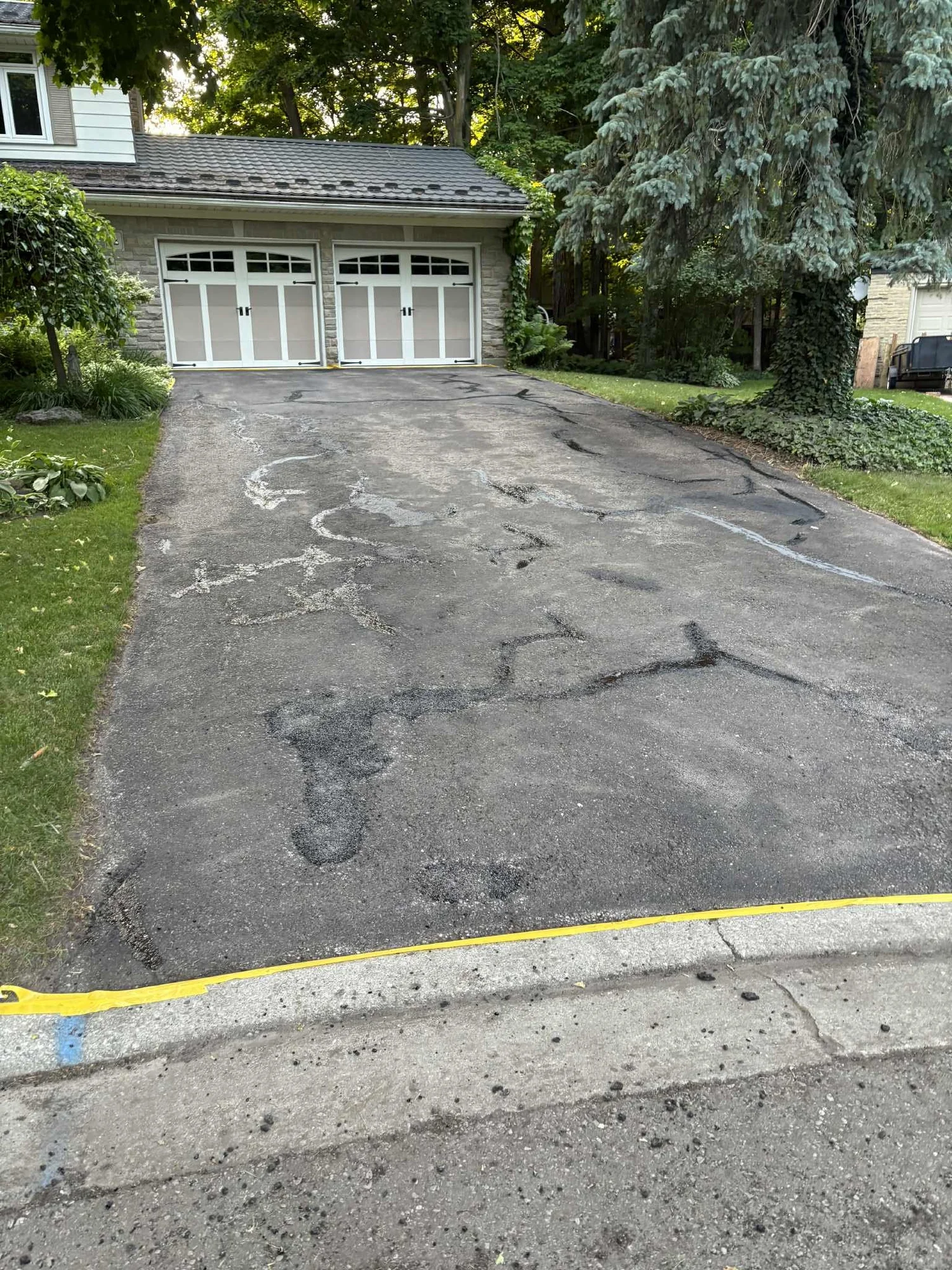 Renewed asphalt driveway leading to a white garage with a pitched roof, surrounded by green grass and trees.