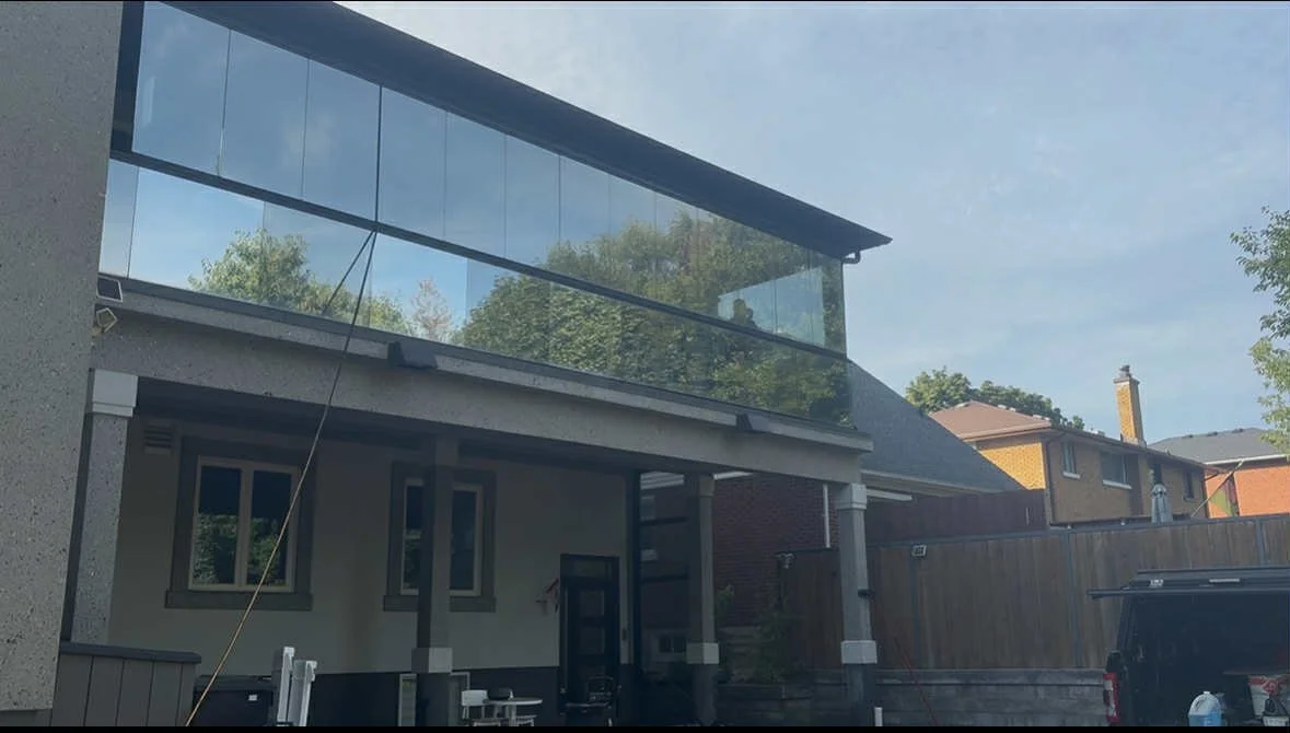 Backyard view of a modern house with a glass enclosed balcony, a covered patio area, and neighboring houses with trees in the background.