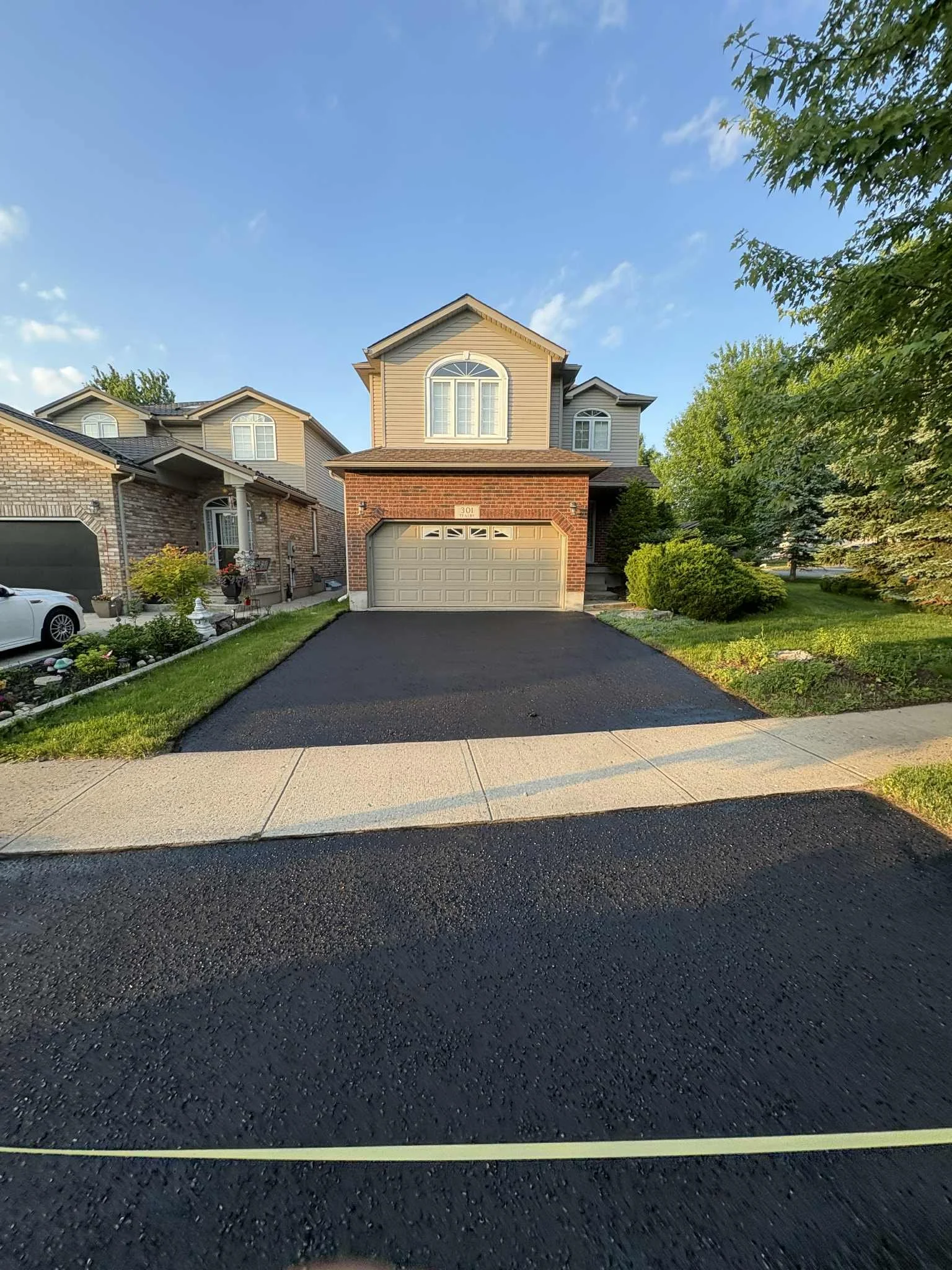 A two-story house with a tan and brick exterior, a gray garage door, and a black asphalt driveway. Green trees and bushes surround the house, and the sky is blue with a few clouds.