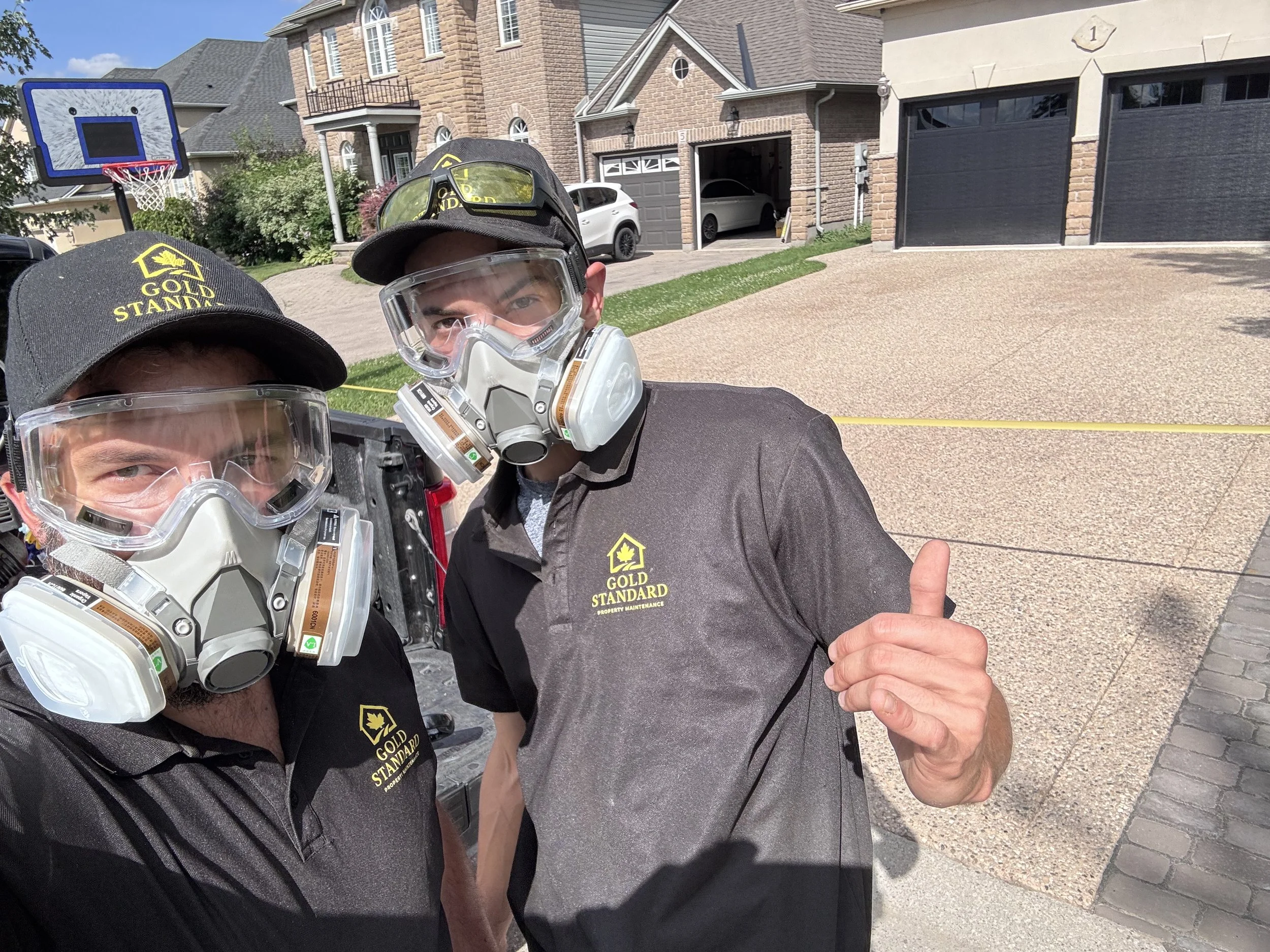 Two workers wearing black uniforms, hats, and safety masks in a residential driveway. One worker's hat and shirt have the words 'Gold Standard' with a logo. They are taking a selfie with a house in the background.