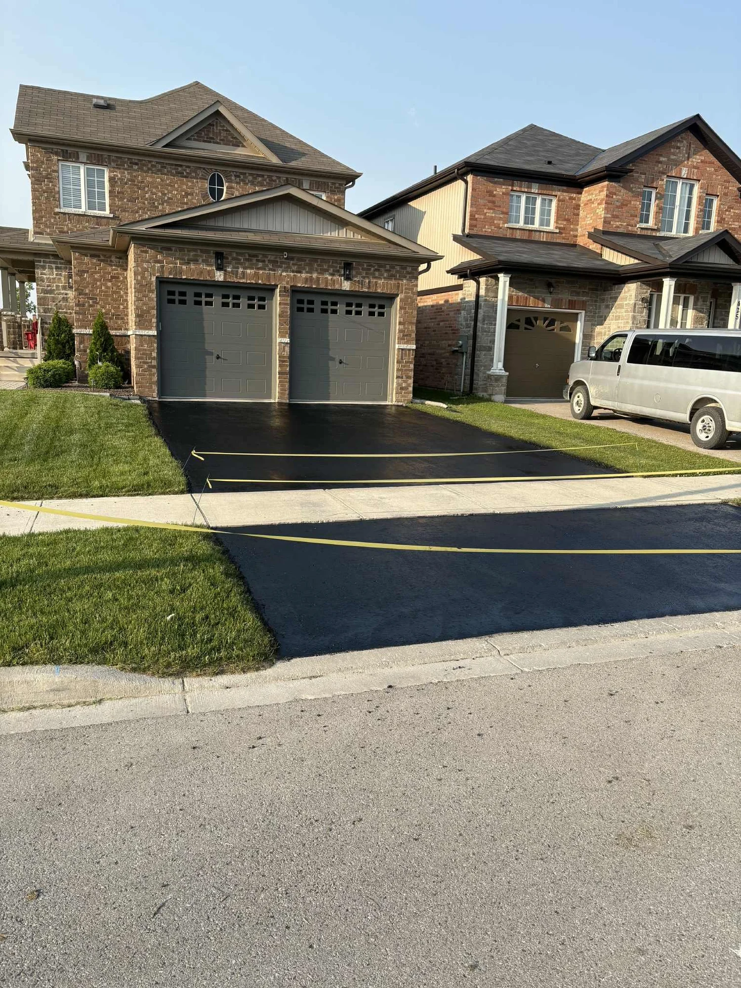 Newly paved black asphalt driveway with yellow caution tape, in front of two houses, including a brick house with twin garage doors, well-manicured lawn, and a white van parked outside.
