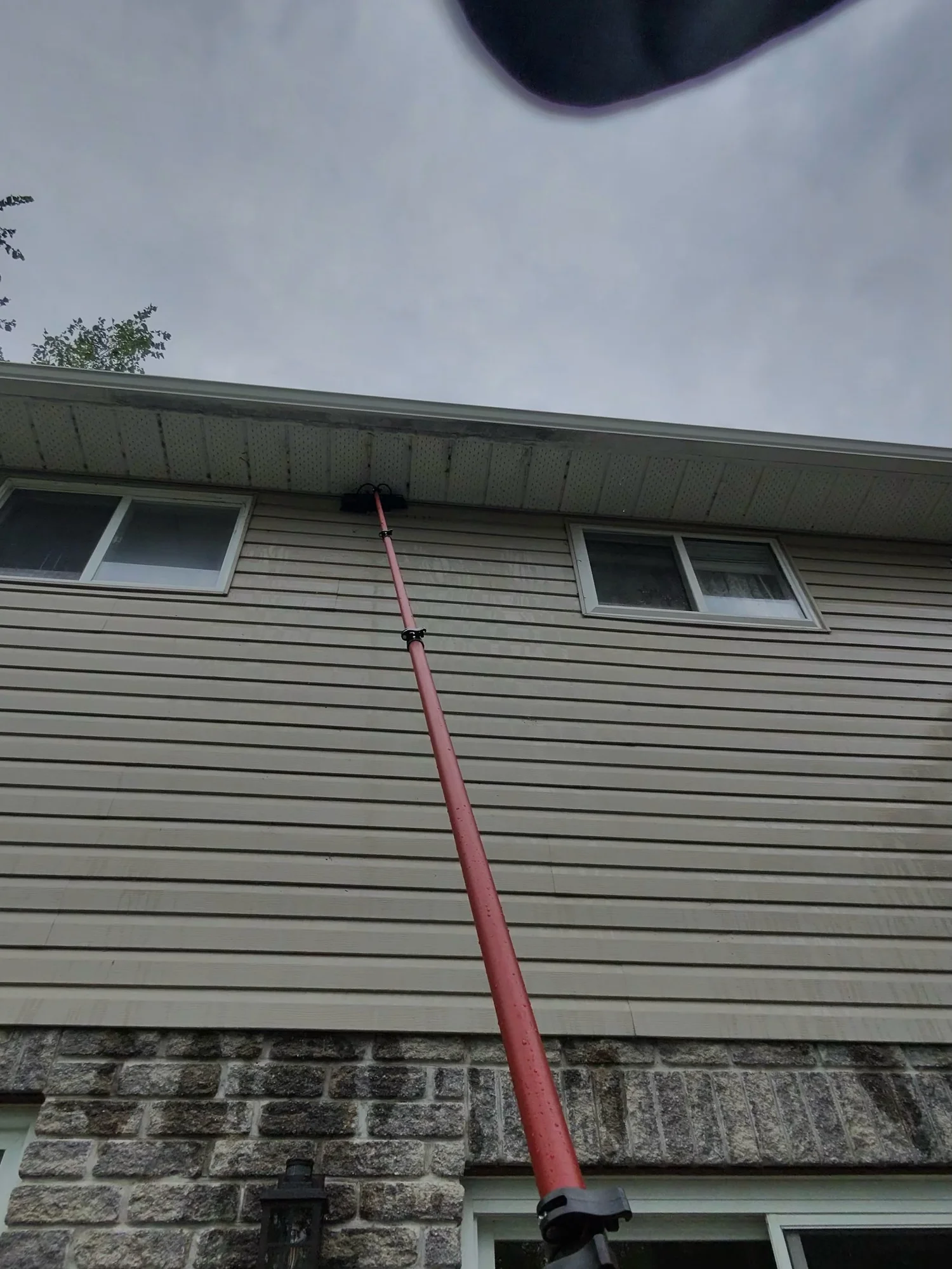A person using a long pole to reach the gutter of a two-story house with beige siding and stone accents, during overcast weather.