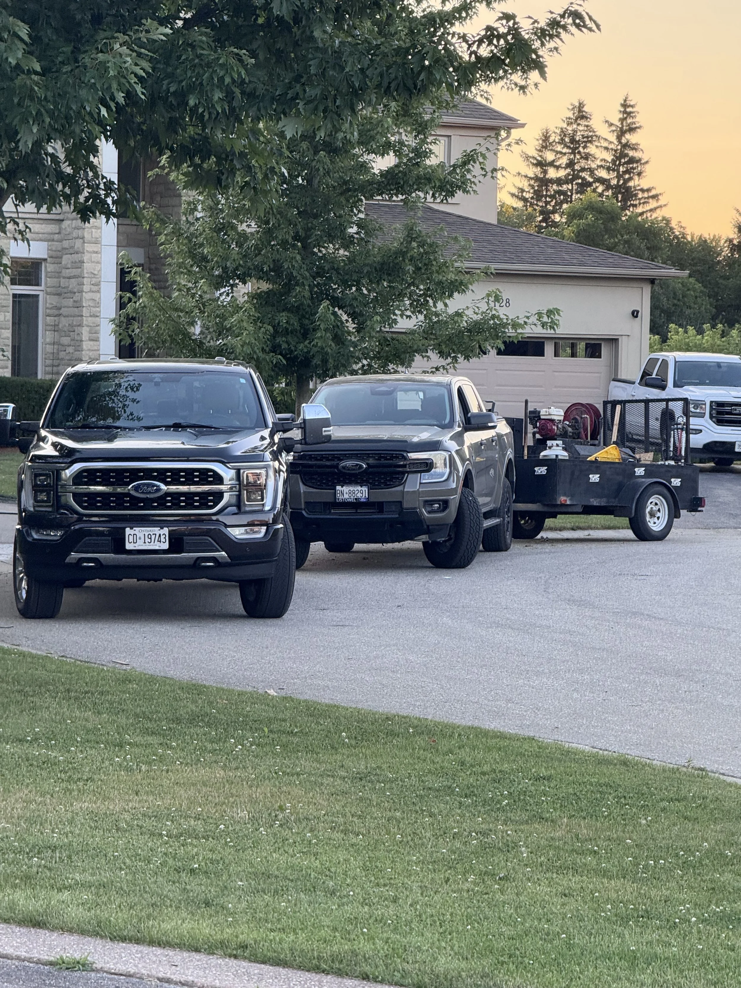 Two pickup trucks parked in front of a house, with a trailer attached to one, during sunset. The house has a two-car garage, and there are trees and a grassy lawn in the foreground.